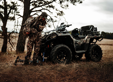 A hunter in camouflage gear adjusts their bow next to a camouflage Polaris Sportsman ATV in a dry, grassy field near a tree.
