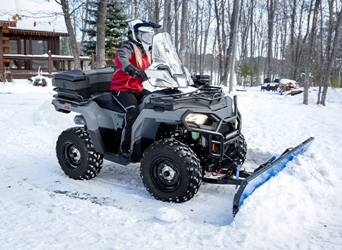 A person in a helmet and winter gear uses a Polaris Sportsman ATV with a snowplow attachment to clear snow from a driveway near a cabin.