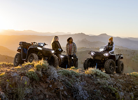 Three people with two Polaris Sportsman ATVs on a rocky hilltop at sunset, overlooking a mountain range.