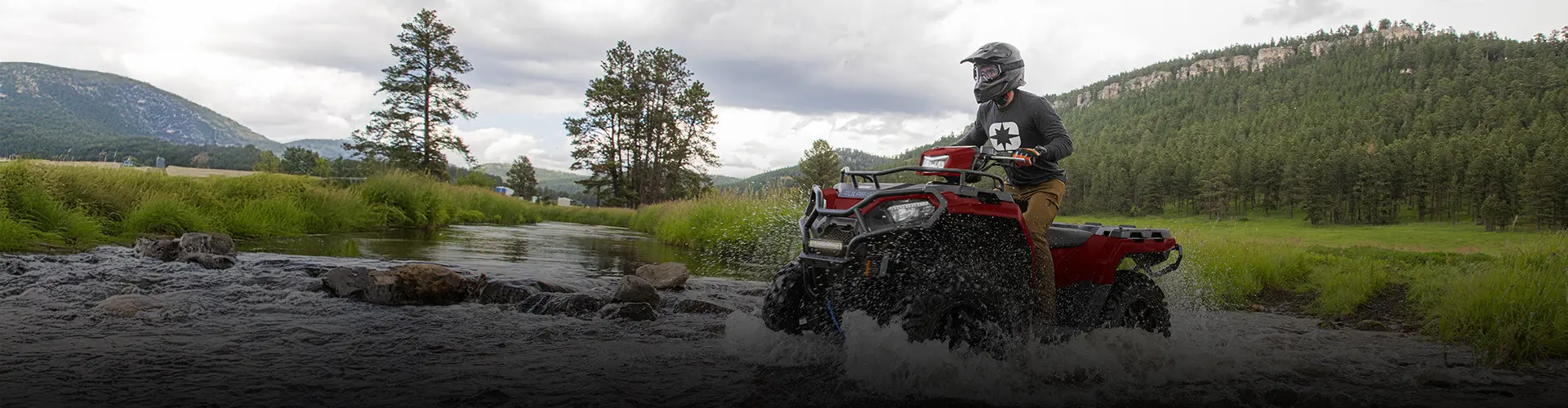 A man on a red Polaris Sportsman ATV rides through a rocky stream, splashing water.