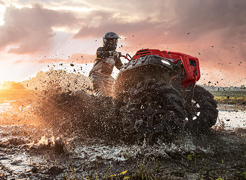 A person on a red Polaris Sportsman ATV speeds through deep mud, kicking up a large splash, with a vibrant sunset in the background.