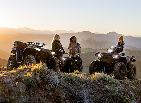 Three people with three Polaris Sportsman ATVs are gathered on a rocky, brush-covered mountain overlook at sunset, with a vast mountain range in the background.