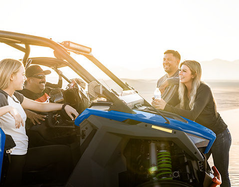 Four people interacting around a blue Polaris off-road vehicle in a desert-like environment.