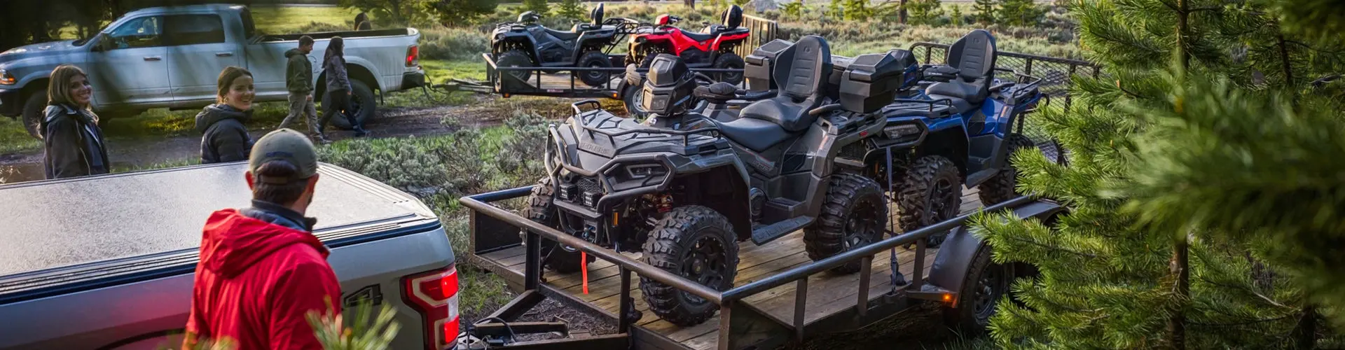 A group of people gathered around Sportsman ATVs loaded onto trailers behind pickup trucks.