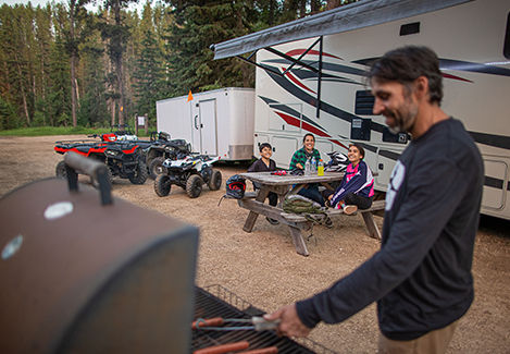 An enclosed trailer parked at a campsite, used for hauling Polaris Sportsman and Youth ATVs, with a family nearby.