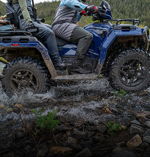 ATV Polaris Sportsman azul con dos conductores salpicando agua y lodo en un área arbolada.