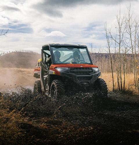 Two people in a Polaris Ranger XP 1000 side-by-side driving on a muddy off-road trail.