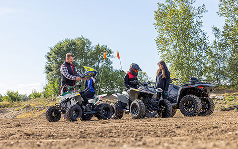 A father helps his daugther with their helmet while they sit on a Polaris Outlaw 110 EFI ATV. The mother stands next to another ATV in a wooded area with her son sitting on a Polaris Phoenix 200 ATV.