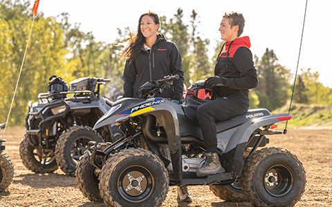 A woman smiling while standing near a teenager seated on a Polaris Phoenix 200 youth ATV.