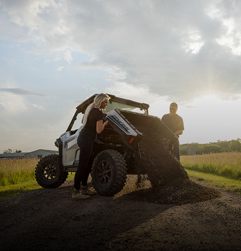 Couple with a Polaris GENERAL XP 1000 UTV loaded with dirt in a sunny outdoor field.