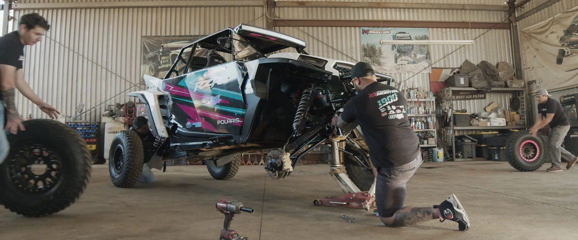 Fernanda Montiel's Polaris RZR SxS undergoing maintenance in a garage, with mechanics changing tires and working on the suspension.