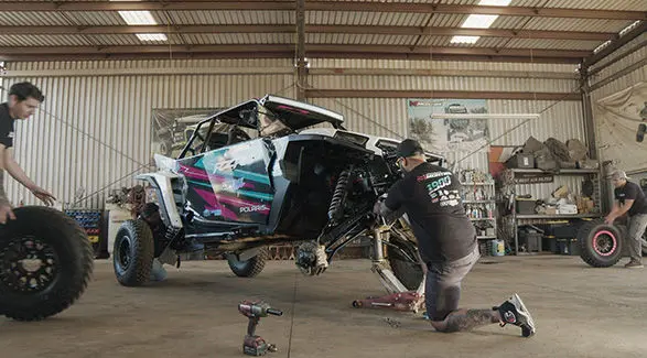 Fernanda Montiel's Polaris RZR SxS undergoing maintenance in a garage, with mechanics changing tires and working on the suspension.