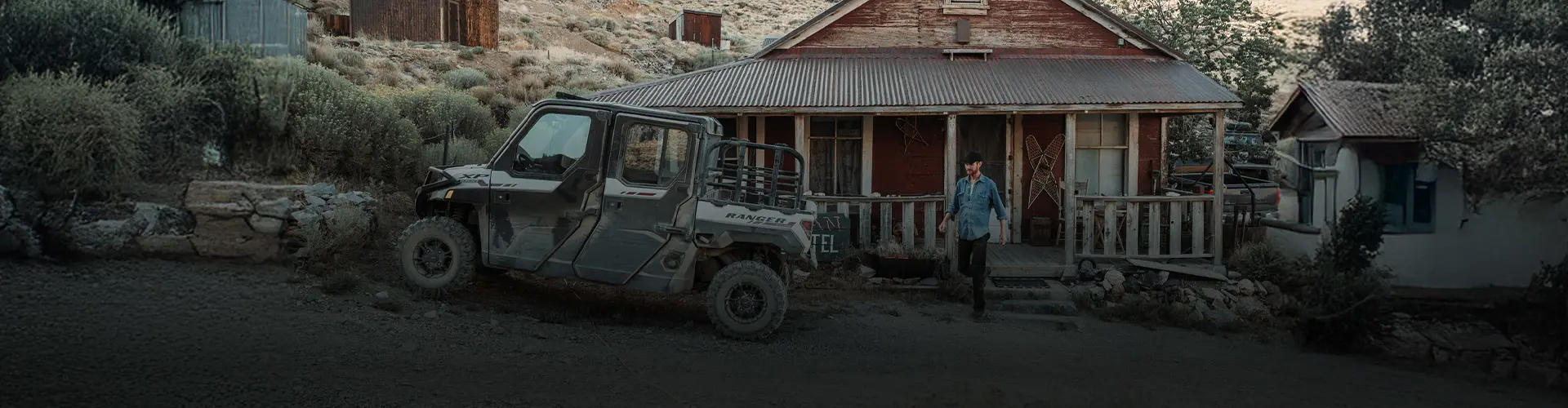 Brent Underwood walks away from a rustic red house with a Polaris RANGER XP 1000 parked on a dirt path beside it, at the Cerro Gordo old town setting.