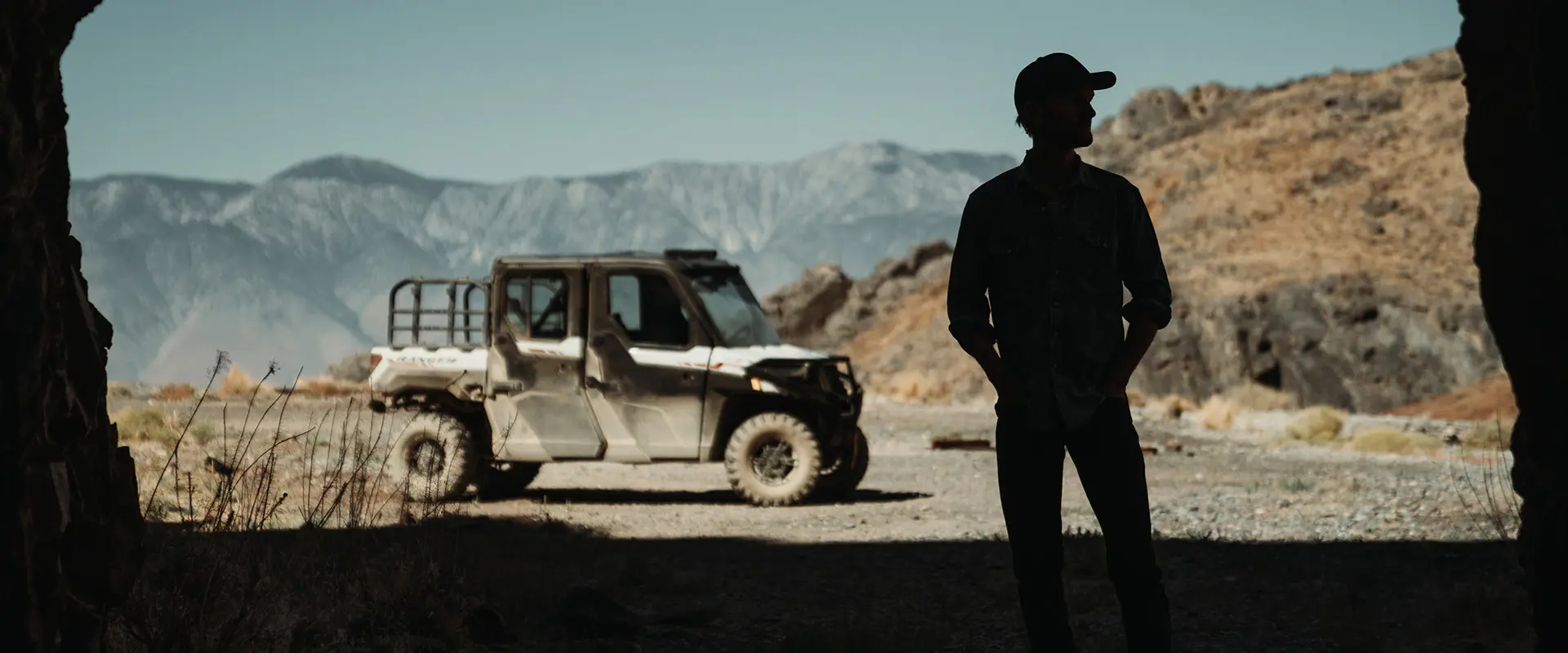 Silhouette of Brent Underwood standing in a cave entrance, looking towards a Polaris RANGER XP 1000 and mountains under a clear sky.