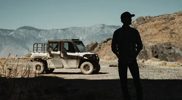 Silhouette of Brent Underwood standing in a cave entrance, looking towards a Polaris RANGER XP 1000 and mountains under a clear sky.