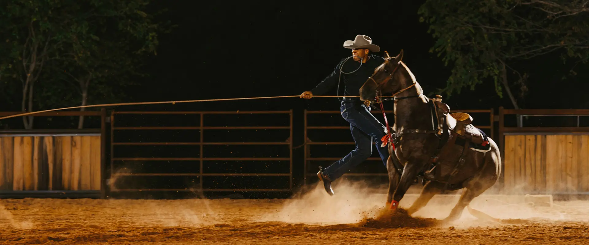 Shad Mayfield, a cowboy on horseback, actively throws a rope in a dimly lit arena, with dust rising from the horse's hooves.