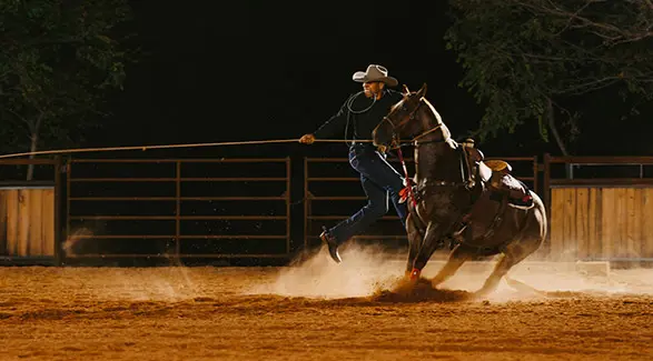 Shad Mayfield, a cowboy on horseback, actively throws a rope in a dimly lit arena, with dust rising from the horse's hooves.