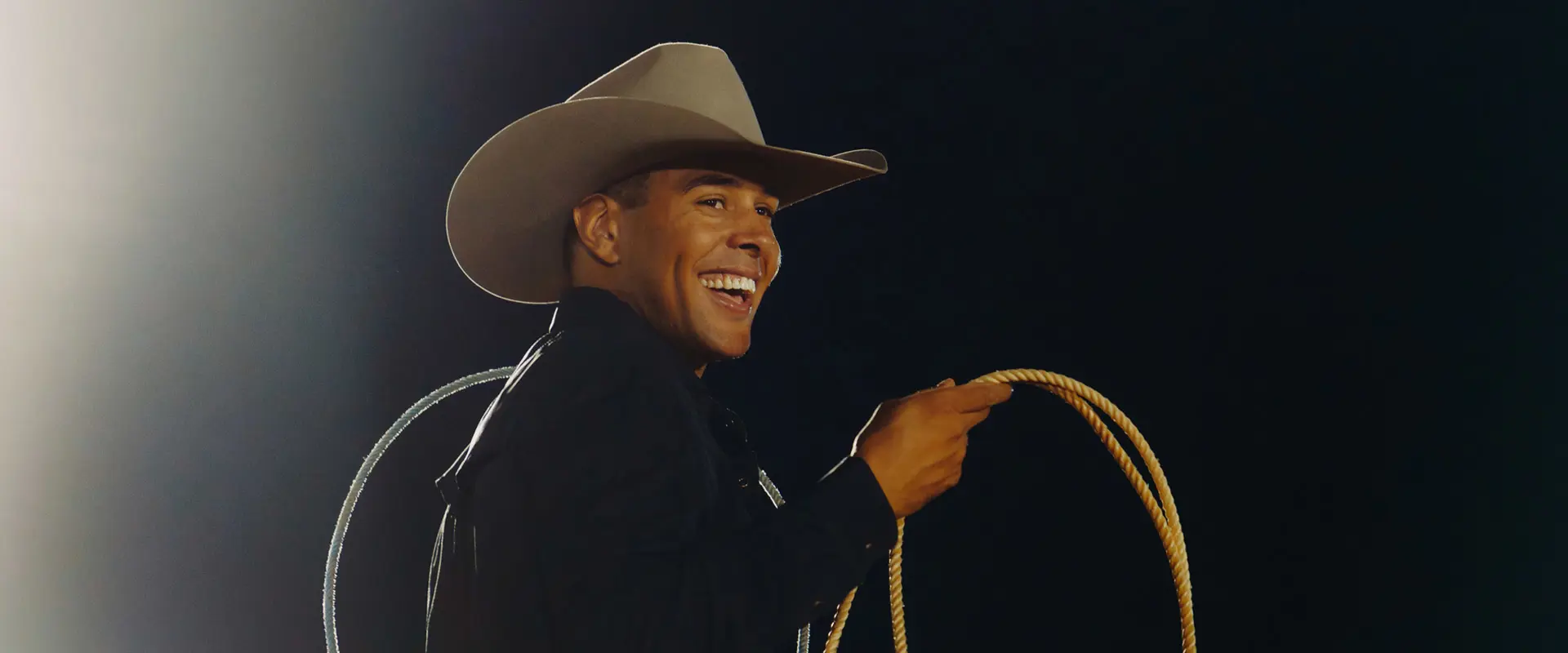 A smiling cowboy, Shad Mayfield, holding a coiled rope, illuminated by a spotlight against a dark background.