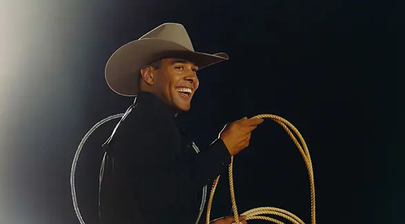 A smiling cowboy, Shad Mayfield, holding a coiled rope, illuminated by a spotlight against a dark background.