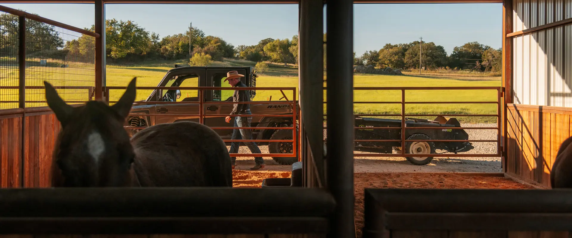 Seen from inside a horse stall, Shad Mayfield walks beside a Polaris RANGER XD 1500 NorthStar Edition UTV that is towing a trailer, with green fields in the background.