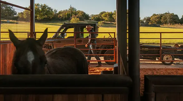 Seen from inside a horse stall, Shad Mayfield walks beside a Polaris RANGER XD 1500 NorthStar Edition UTV that is towing a trailer, with green fields in the background.