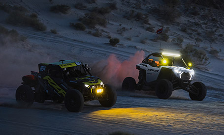 Two Polaris RZR SxS racing across a desert at night, with bright LED headlights and light bars cutting through the dark.