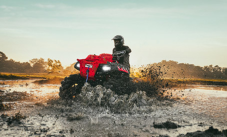 A red Polaris Sportsman High Lifter ATV creates a large splash while driving through a muddy field at sunset.