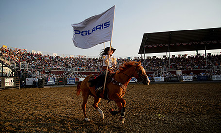 Rider on horseback carrying a large Polaris flag across a rodeo arena, with packed grandstands and event signage visible in the background.