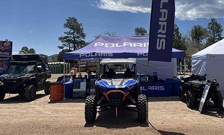 Polaris RZR Side-by-Side displayed in front of a Polaris-branded event tent, with flags, promotional displays, and additional off-road vehicles at an outdoor expo.