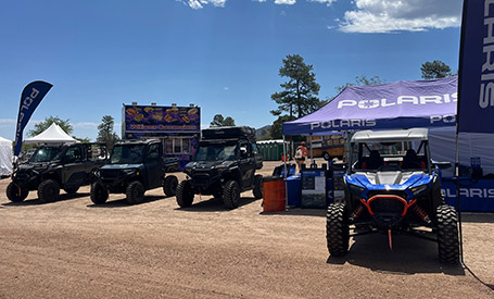 Polaris RZR Side-by-Side displayed in front of a Polaris-branded event tent, with multiple Polaris RANGER UTVs parked nearby at an outdoor expo under a bright sky.