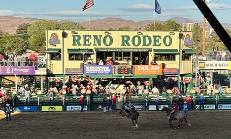 Wide view of the Reno Rodeo arena with riders on horseback competing in front of packed grandstands, sponsor signage, and the large Reno Rodeo display board.