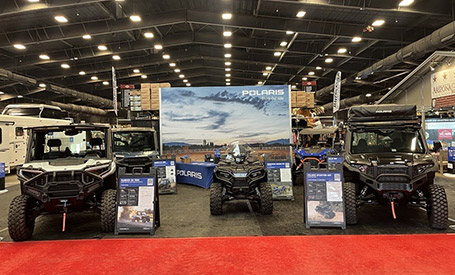 Polaris trade show booth featuring a white Ranger UTV, a black Sportsman ATV, and a black XPEDITION UTV displayed on a red carpet.