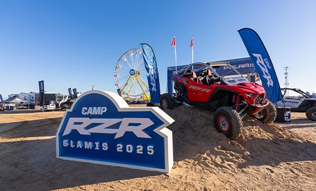 Camp RZR display with red Polaris side-by-side vehicle on sand dunes at Glamis 2025 event under clear blue sky.