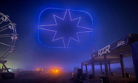 Blue illuminated Polaris RZR star-shaped drone light display above foggy event grounds at night with Ferris wheel in background.