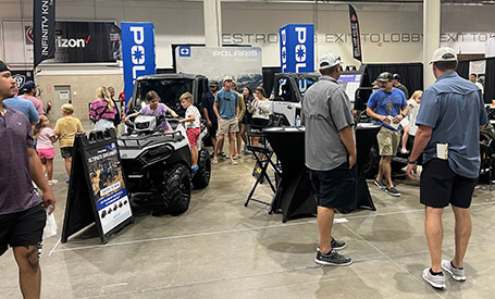 Polaris Sportsman ATV and RANGER UTVs displayed indoors at a Polaris event booth, with attendees gathered around product displays, and promotional setups.