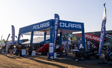 Large Polaris-branded outdoor event booth featuring Polaris RZR SxS vehicles, product displays, flags, and attendees gathered around multiple vendor setups.