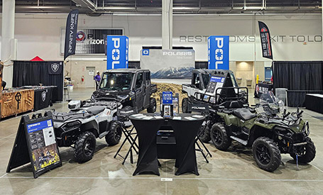 Polaris Sportsman ATVs and RANGERS UTVs displayed indoors at a Polaris event booth, with attendees gathered around product displays, and promotional setups.
