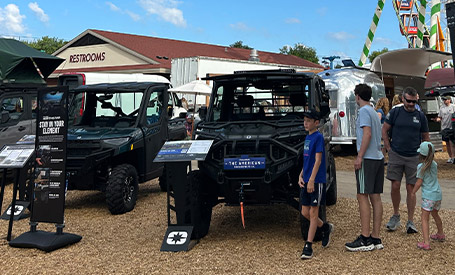 Polaris event booth with multiple Polaris RZR and Polaris GENERAL SxS vehicles displayed under a large blue branded tent, with attendees walking around on a sunny day.