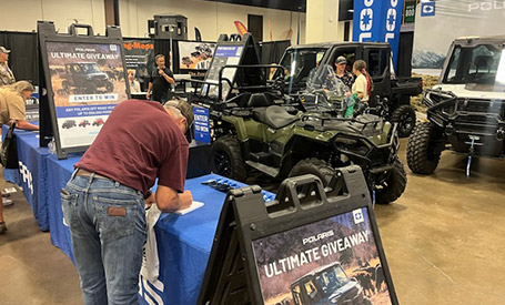 Polaris RANGER UTV and Polaris Sportsman ATV displayed indoors at a Polaris expo booth, with giveaway signage, branded banners, and attendees.