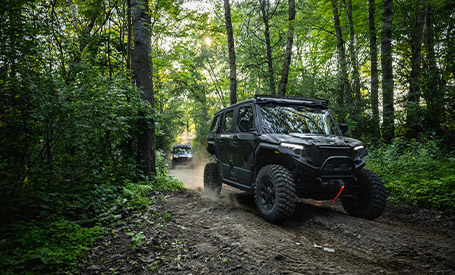 Polaris XPEDITION ADV SxS driving along a wooded trail, kicking up dust while navigating a narrow forest path with another vehicle following behind.