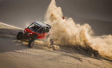 A red Polaris RZR Pro R SxS speeding down a desert sand dune, kicking up a massive plume of sand.