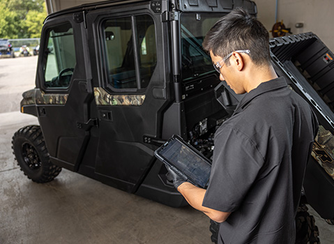 A Polaris technician in safety glasses uses a tablet while standing next to a black and camouflage Polaris RANGER CREW XP UTV with its cargo bed tilted up.