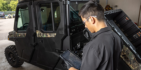 A Polaris technician in safety glasses uses a tablet while standing next to a black and camouflage Polaris RANGER CREW XP UTV with its cargo bed tilted up.