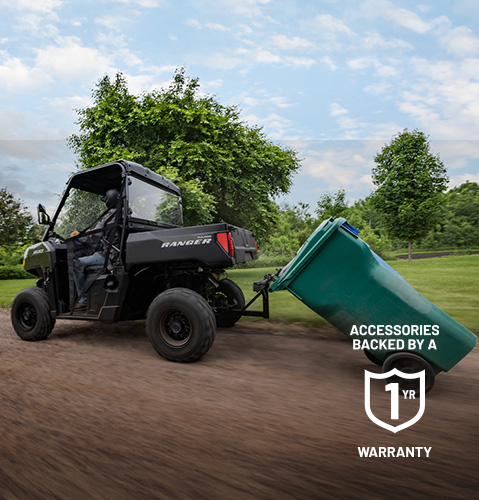 A Polaris RANGER 500 UTV drives along a dirt path, towing a large green trash can through a grassy area with trees.