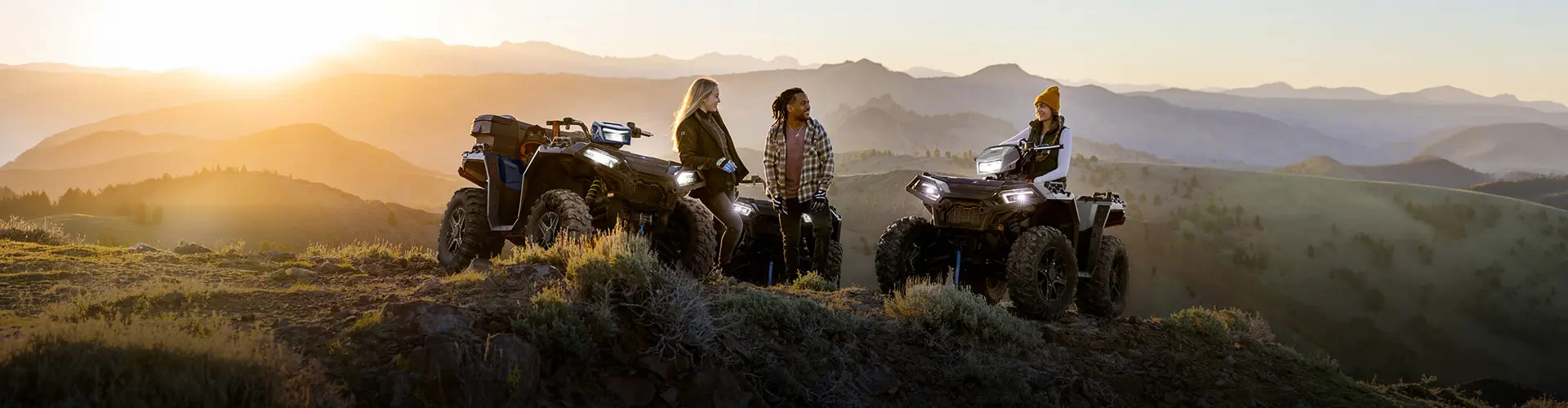 Two Polaris Sportsman ATVs on a dirt road at an overlook, with a family around and sitting on them and looking at a vast forest valley.