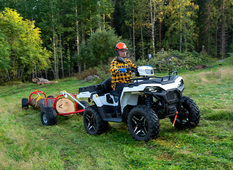 Polaris Sportsman ATV towing logs across a grassy field, demonstrating utility and work capabilities in a wooded setting.