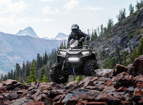 Polaris Sportsman ATV navigating a rocky mountain trail, showcasing trail-riding capability with alpine peaks and forested slopes in the background.