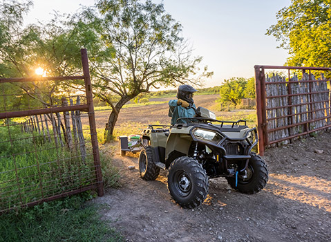 Polaris Sportsman ATV towing equipment through a ranch gate at sunset, highlighting work and property maintenance capabilities in a rural setting.