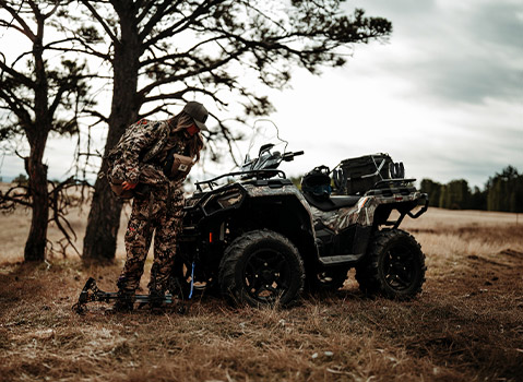 Polaris Sportsman ATV in camouflage parked in a field beside a hunter with archery equipment, highlighting hunting and outdoor recreation capabilities.