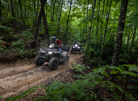 Two Polaris Sportsman ATV riders traveling along a wooded trail, demonstrating recreational riding through dense forest terrain.